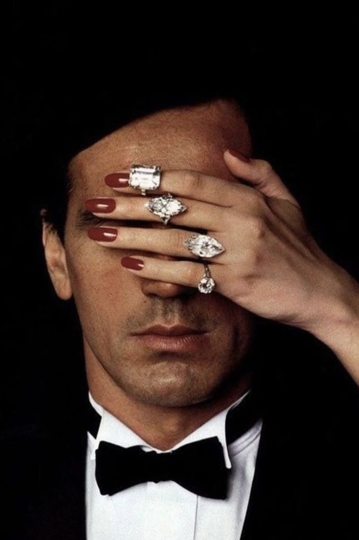 A close-up portrait of a man in a black tuxedo and bow tie, with a hand covering his eyes. The hand is adorned with multiple large, prominent silver rings featuring clear gemstones.