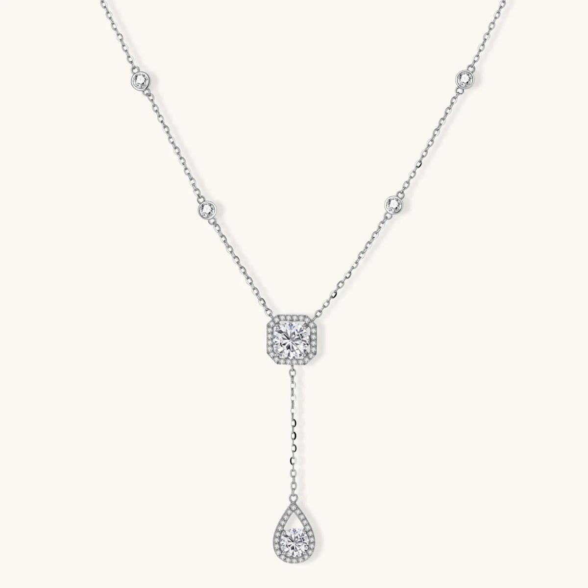A delicate silver-toned Y-necklace featuring a square halo pendant and a teardrop-shaped drop pendant, both adorned with small clear stones, set against a solid off-white background.